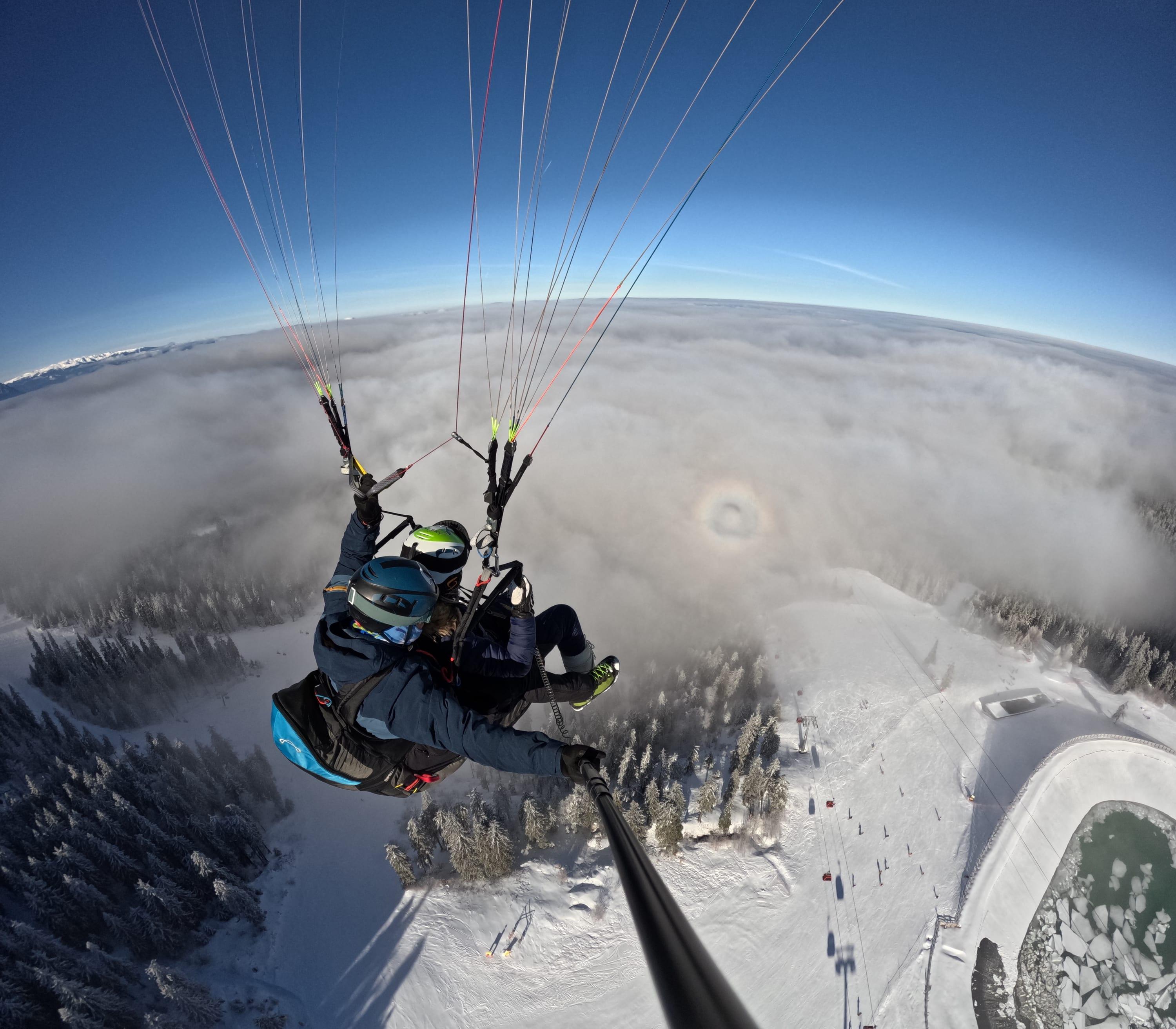 Pilot și pasager în tandem deasupra mării de nori, cu pârtia înzăpezită dedesubt
